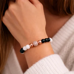 Női ásvány karkötő csuklón viselve, fehér és fekete gyöngyökkel, rozéarany díszítőelemekkel és csillogó központi gyönggyel